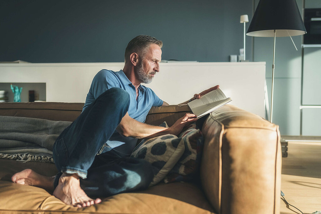 Mann in Wohnung sitzt auf Couch und liest ein Buch