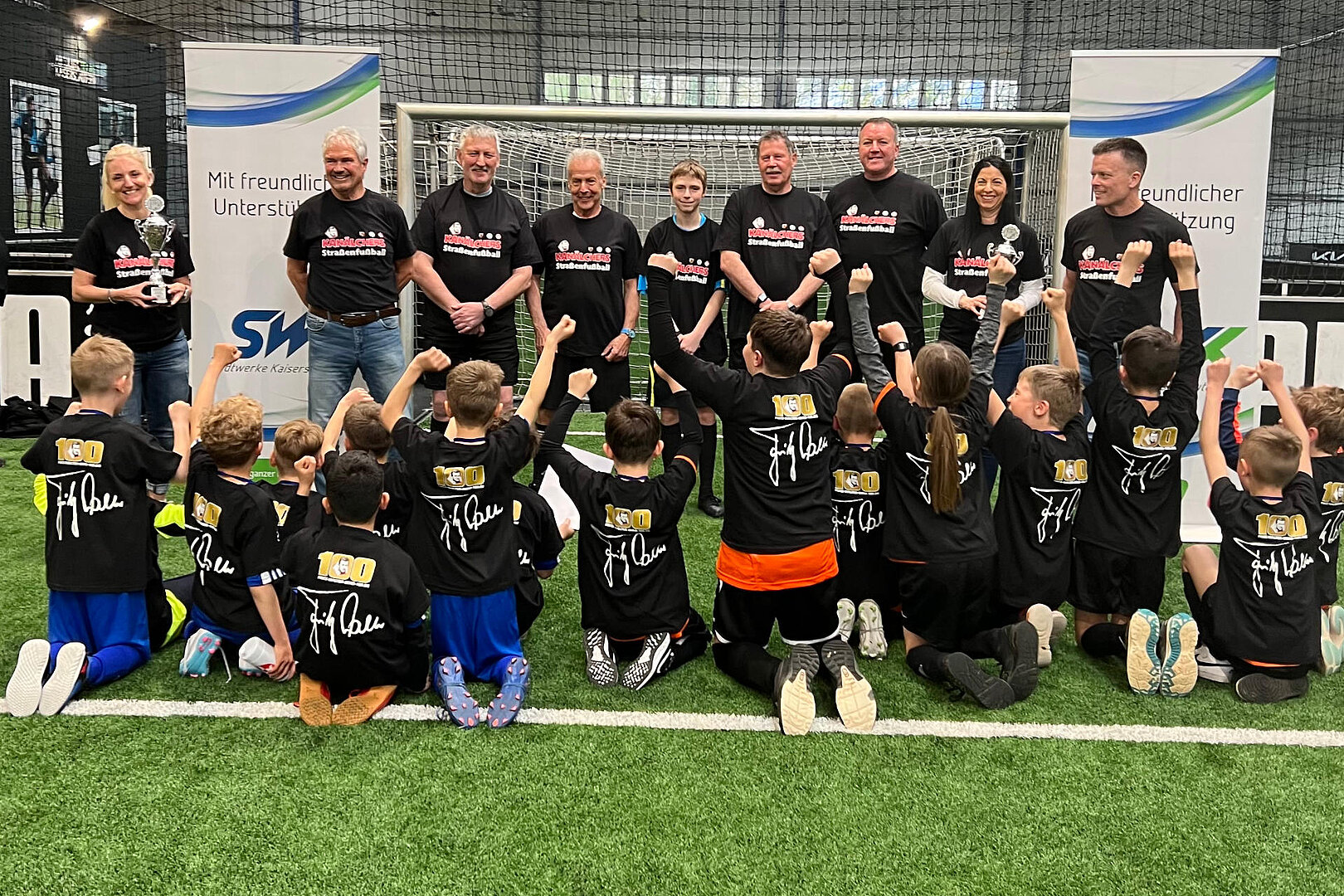 Fußball-Halle auf dem Rasen jubeln Kinder, vor den Kindern stehen Personen in einer Reihe und eine Person von SWK überreicht einen Pokal zur Fußballstadtmeisterschaft