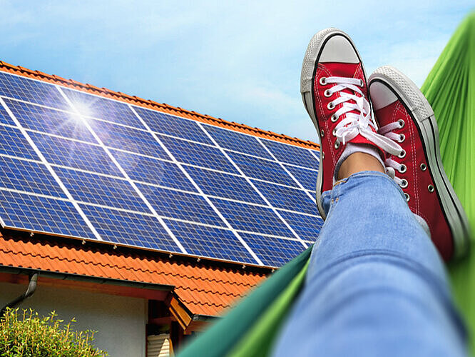 Beine und Füße einer Person auf Hängematte mit Blick auf Haus mit Solaranlage