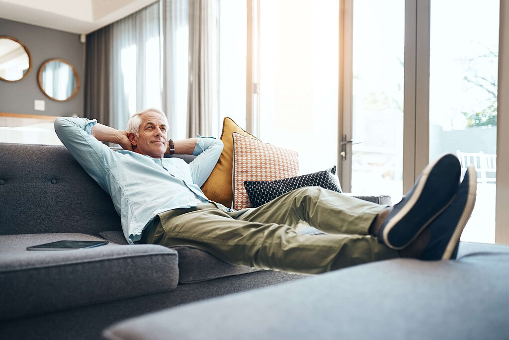 Mann auf Couch neben sich ein Tablet und legt die Beine hoch, verschränkt die Arme hinter Kopf