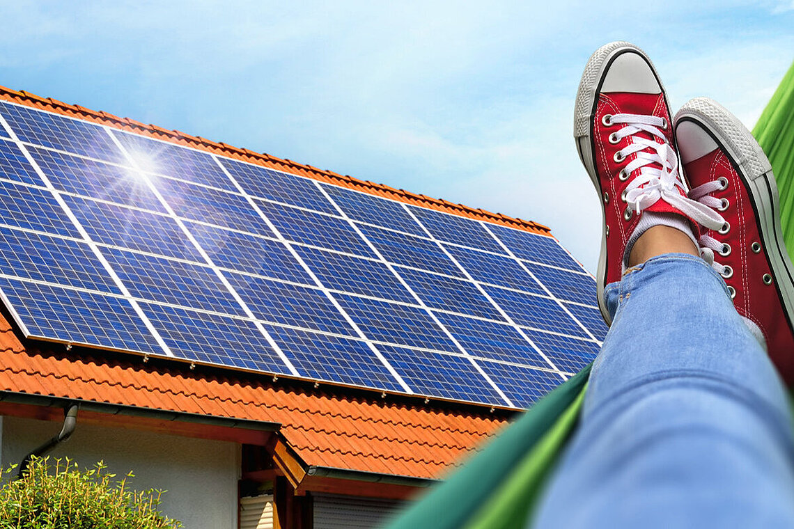 Beine und Füße einer Person auf Hängematte mit Blick auf Haus mit Solaranlage