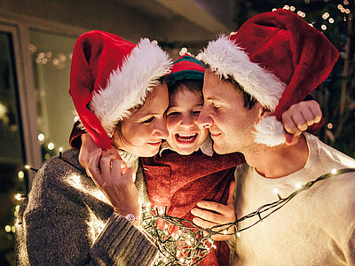 Mann, Frau und Kind mit Weihnachtsmützen und Lichterketten umhängen