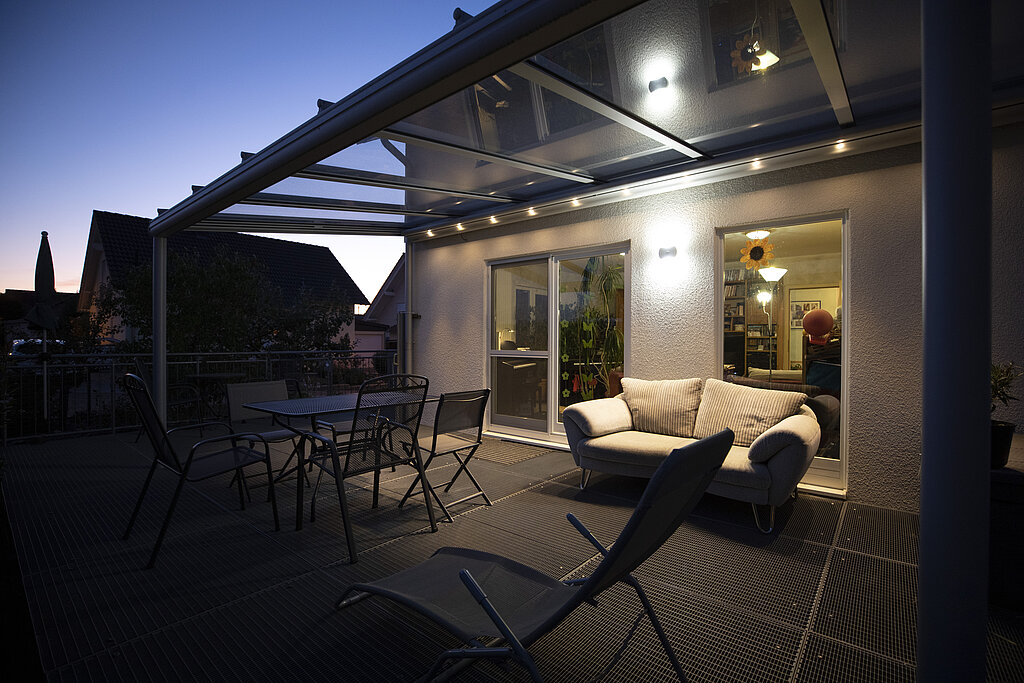 Veranda mit Glasvordach Tischen und Stühlen bei Sonnenuntergang mit Einblick in die Wohnung